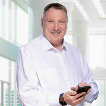 Portrait von Volker Rozek. Er steht in einer Büroumgebung, trägt ein weßes Hemd und hält ein Handy in der Hand